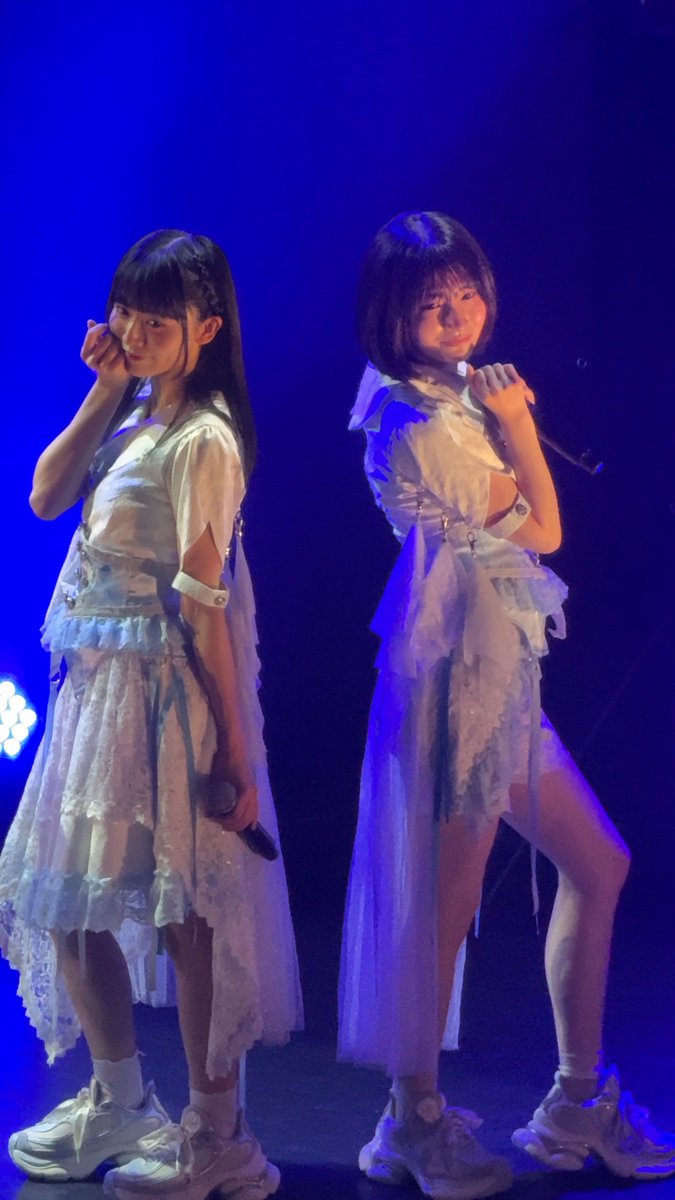 Two young female performers on stage under blue lighting, wearing matching white frilly outfits and chunky white sneakers while holding microphones.