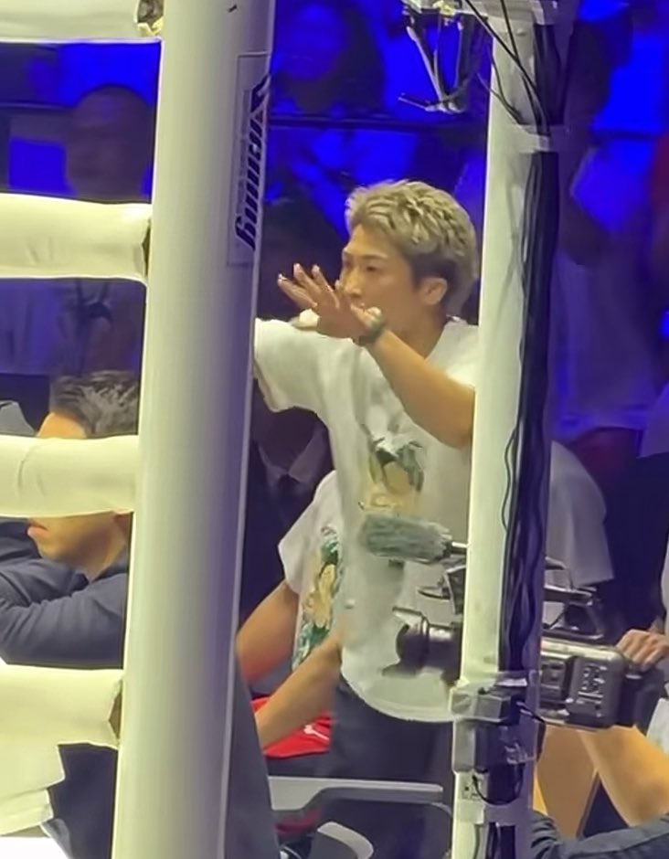 A young man with blonde hair wearing a white graphic t-shirt appears to be backstage or in a filming area, gesturing with his hand near his face next to some equipment and rigging.