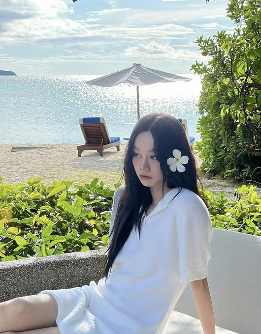 A woman with long black hair and a white plumeria flower behind her ear sits in a white outfit at a tropical beach resort, with the ocean and umbrella visible behind her.