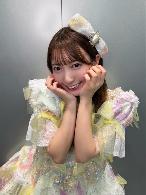 A woman wearing an elaborate, pastel-colored frilly outfit with a matching bow poses with her hands framing her face and smiling.