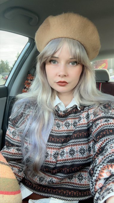A selfie of a young woman with long silver/grey hair, wearing a tan beret and a patterned sweater with red, white, and black designs, sitting inside a car.