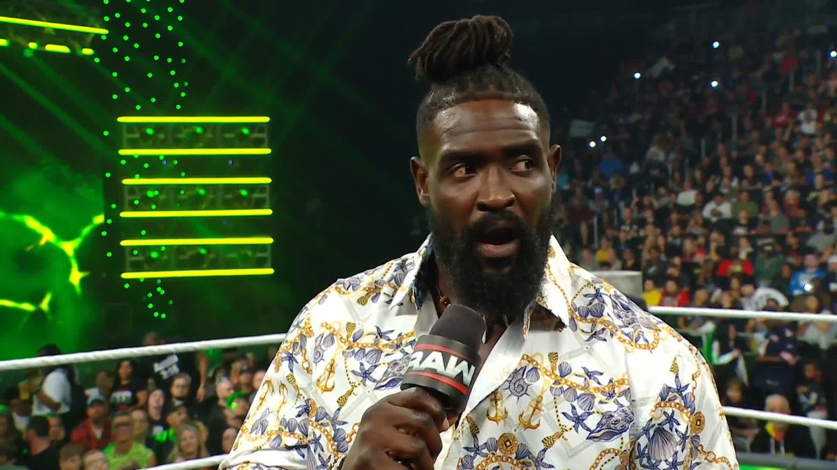 WWE wrestler Oba Femi, identified by the post's "@Obaofwwe" tag, stands in a wrestling ring holding a "RAW" branded microphone while addressing a crowd, wearing a patterned white shirt with gold and blue nautical designs. His raised eyebrows and wide-eyed expression align with the captioned line "I'M THE CAPTAIN NOW," suggesting he is delivering a confident in-ring promo announcing his leadership role.