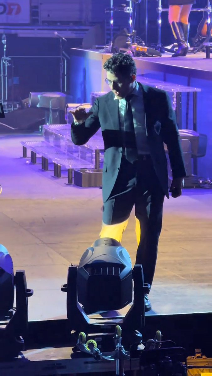 A man in a black suit and sunglasses stands on a stage illuminated by blue lighting, captured mid-gesture during what appears to be a concert.