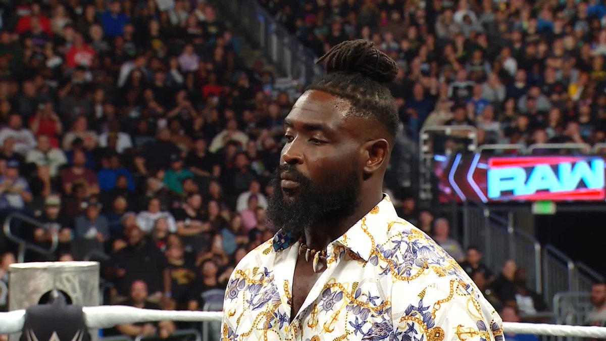 A man with a beard and top knot, wearing a white and gold Versace-style patterned shirt and a bone necklace, stands in a wrestling ring with the "RAW" logo visible on the jumbotron behind him, fitting the #WWERaw setting announced in the post.