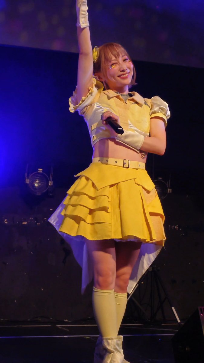 A performer in a yellow ruffled crop top and skirt with knee-high boots and gloves waves to the audience while holding a microphone on a blue-lit stage.