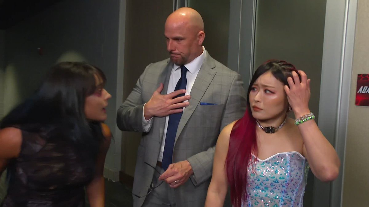 A backstage scene shows a bald man in a gray suit standing between two women, one with dark hair and another with pink-streaked hair wearing a sparkly corset dress, appearing to be in conversation.