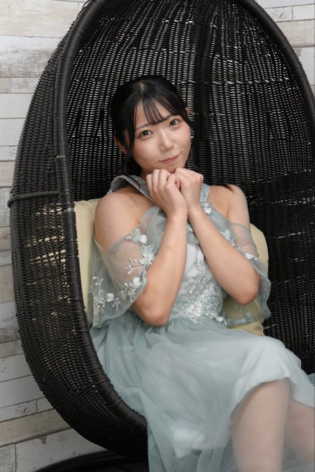 A young woman with dark hair sits in a black wicker hanging chair, wearing a light blue and silver floral dress with sheer sleeves, resting her chin on her hands.