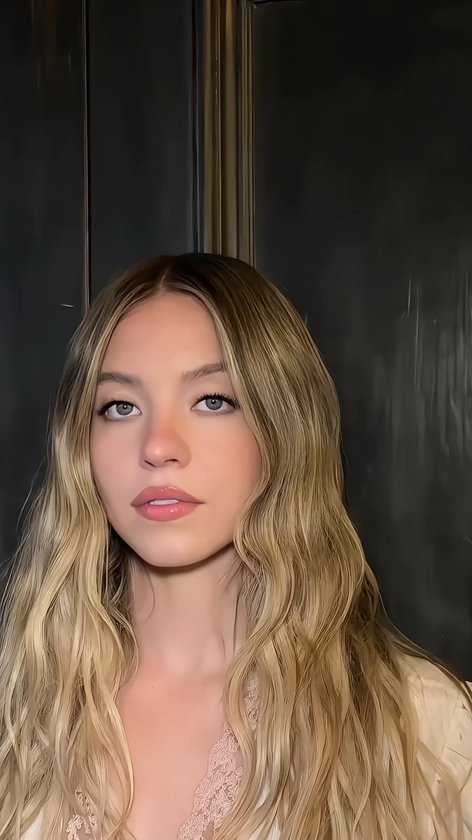 A close-up portrait of a young woman with long, wavy blonde hair, light eyes, and glossy makeup in a soft-lit setting.