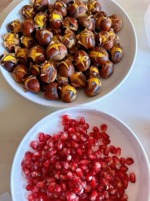 Two white bowls on a table - the top bowl filled with roasted chestnuts that have been scored and partially peeled, and the bottom bowl filled with bright red pomegranate seeds.
