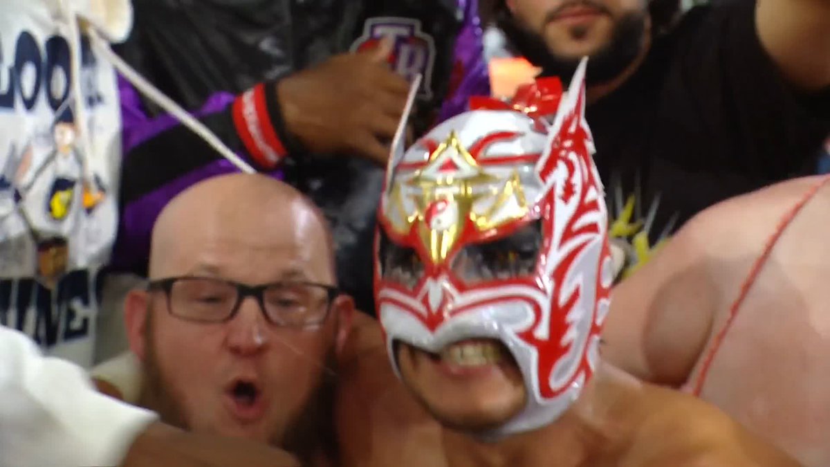 Two enthusiastic wrestling fans react with open-mouthed shock, one wearing a distinctive red and silver lucha libre-style mask, as they witness Dragon Lee defeating Big Cass.