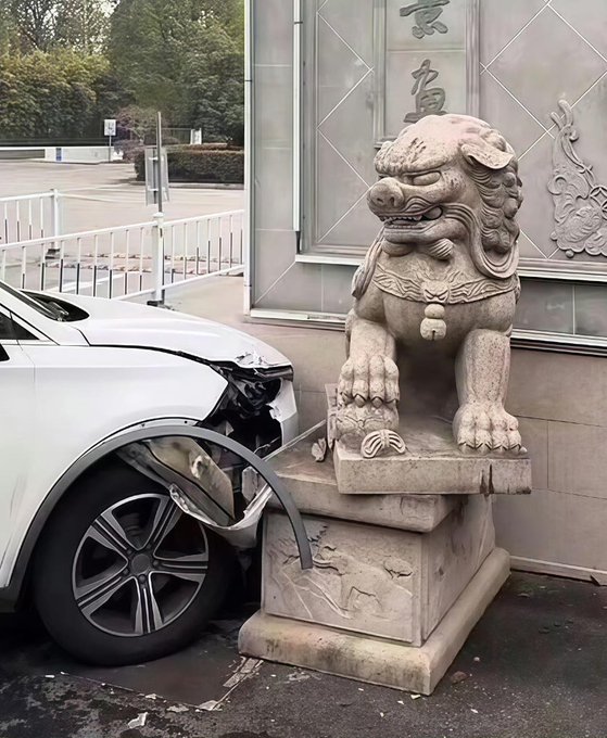 A white car has crashed into a stone Chinese guardian lion statue at the entrance of a building, with visible damage to the car's front end.