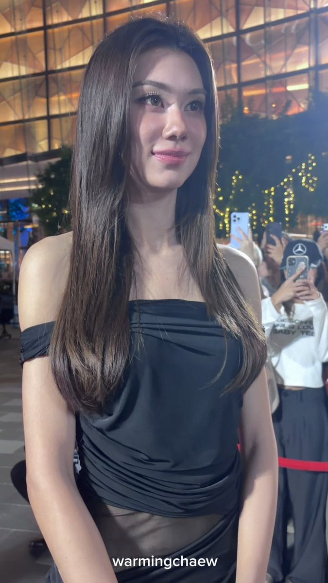 A woman with long brown hair wearing a black off-shoulder top poses at what appears to be a glamorous event venue with decorative lights, while fans in the background take photos with their phones. The watermark "warmingchaew" appears at the bottom of the image, suggesting a celebrity encounter that aligns with the post's theme of unexpectedly running into someone before a trip to Hong Kong.