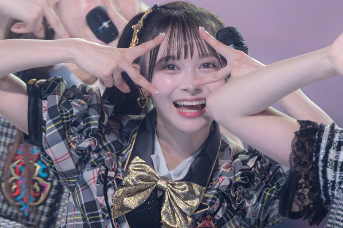 A young woman with bangs performs on stage, making a peace sign with both hands while wearing a plaid outfit with a gold bow.