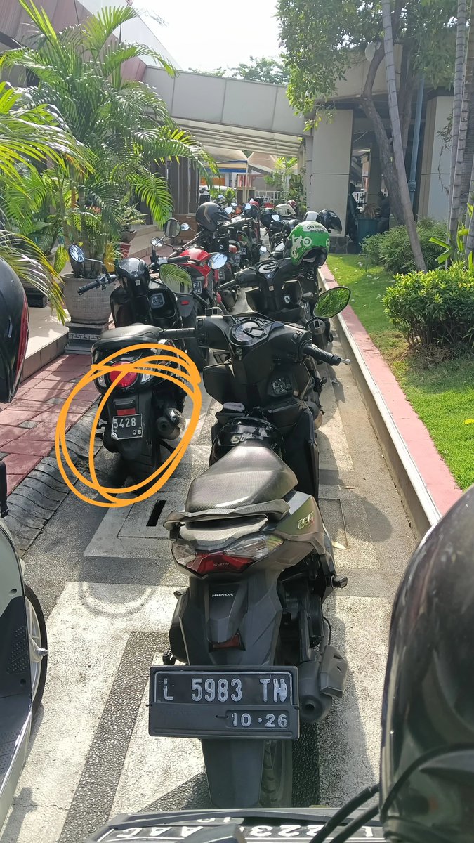 Smash1014314's tweet photo. Mari kita bahas dugaan pungli di Samsat Manyar Surabaya buat kalian yang mau perpanjang plat nomor 5 tahunan.

Kalau plat kendaraan kalian rusak atau bahkan tinggal secuil bagian besinya, mungkin sebaiknya dilengkapin dulu. 😭

Kronologinya begini.

Gue datang ke Samsat Manyar buat mutasi keluar sekaligus perpanjang plat. Kebetulan plat belakang gue hilang waktu berangkat kerja di Gresik. Semua surat-surat lengkap.

Pas antre cek fisik, awalnya aman-aman aja. Petugasnya juga melakukan gosok nomor rangka/mesin seperti biasa.

Setelah selesai, orang yang ngecek malah bilang,

“Seikhlase ae, Mas.”

Karena menurut gue itu termasuk pungutan, ya gue nggak kasih.

Pas gue mau pergi, tiba-tiba gue diteriaki, dicegat, terus plat kendaraan gue difoto.

“Plat belakang mana?”

Padahal dari awal dia sudah lihat kalau plat belakang gue memang nggak ada. Gue udah feeling bakal dipersulit gara-gara nggak ngasih uang tadi.

Gue jawab, “Hilang, Pak. Jatuh di jalan waktu berangkat kerja. Udah saya cari lagi tapi nggak ketemu.”

Dari situ gue makin ngerasa bakal ada masalah.

Gue juga sempat tanya ke petugas cek fisik, “Sebelum saya tadi ada orang yang platnya cuma separuh, nggak kelihatan L XXX. Itu bisa lolos, gimana, Pak?”

Dia cuma diem sambil foto kendaraan gue.

Setelah itu gue menuju loket. Sebelum masuk loket, kita diarahkan antre dulu ke dua security buat dicek kelengkapan surat.

Gue tanya ke security soal pungutan tadi.

Jawabannya,

“Emang seikhlasnya aja, Mas.”

Gue tanya lagi, “Kalau nggak dikasih gapapa kan, Pak?”

Dijawab, “Ya lihat aja nanti, Mas, diverifikasi.”

Akhirnya gue tunggu sampai nomor antrean gue dipanggil.

Di loket, surat gue dikasih catatan “plat belakang hilang”. Gue jelasin lagi kondisinya sekaligus nanya soal pungutan tadi.

Petugas loket cuma diem, lalu tanya ke temannya. Temannya malah nyuruh tanya ke loket verifikasi sebelumnya.

Intinya nggak ada yang mau jawab soal pungutan tadi. 😭

Mereka kemudian nyuruh gue ke tukang plat buat pasang plat belakang.

Gue jawab,

“Lah, mending saya ke tukang plat, pasang dua-duanya sekalian, nggak usah bayar pajak ginian.”

Mereka diem.

Gue mikir, buat apa gue harus ke tukang plat dulu, pasang plat lama, balik lagi ke Samsat buat dapet plat baru, terus pasang lagi? Ribet banget.

Gue tanya lagi,

“Memang ada aturannya, Pak? Kalau plat hilang, sudah dicari tapi nggak ketemu, harus bikin baru di tukang plat dulu supaya bisa perpanjang?”

Niat gue sebenarnya cuma mau tahu aturannya, bahkan kalau ada aturan tertulisnya gue mau foto supaya orang lain juga bisa ngerti sebelum datang ke Samsat.

Tapi mereka tetap kekeh nyuruh gue ke bagian verifikasi.

Karena udah kesel, akhirnya gue cabut semua berkas dari map, terus pergi sambil nguntel-nguntel formulir di dalam Samsat. 😭

Akhirnya gue pulang.