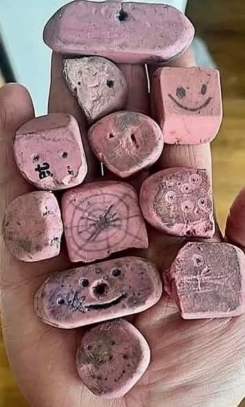A hand holding a collection of pink stones or clay pieces with various carved faces, smileys, and symbols etched into them.
