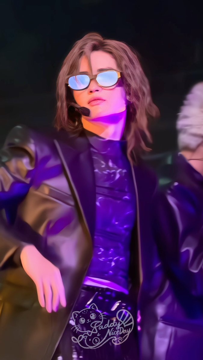 A person with long brown hair wearing blue reflective sunglasses and a black leather jacket, performing on stage under purple lighting, with a watermark reading 'Daddy NiceDay'.