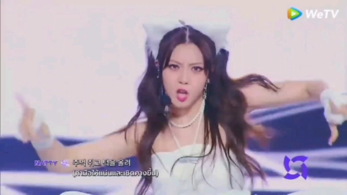 A young female performer with long dark hair and a white bow headband is captured mid-performance on a stage with pink lighting, wearing a white outfit and pearl necklace. The Korean subtitles identify her as "NATTY," with lyrics on screen, and the "WeTV" logo appears in the top right corner alongside a stage logo in the bottom right, matching the post's praise of her stage-ready singing and dancing talents.