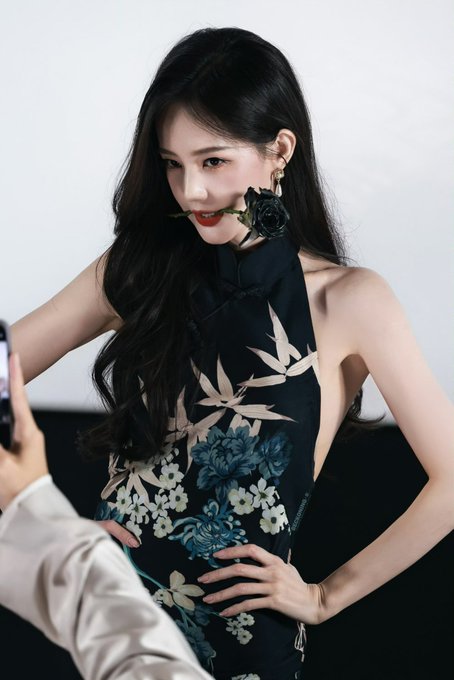 A young woman with long dark wavy hair poses holding a black rose near her face, wearing an elegant black halter cheongsam-style dress adorned with blue and cream floral embroidery, likely backstage or during a photoshoot as suggested by the hand holding a phone in the foreground.