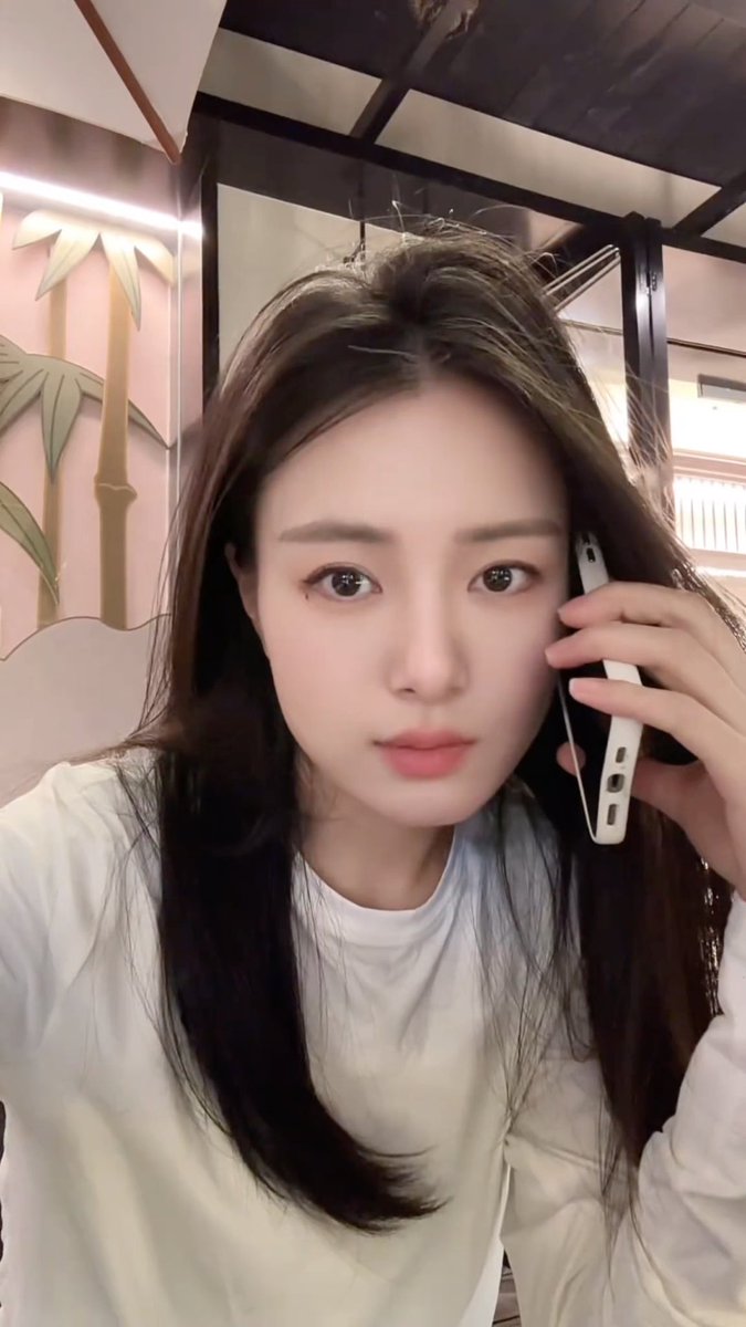 A young woman with long dark hair takes a selfie while holding a white smartphone to her ear, in what appears to be a restaurant setting.