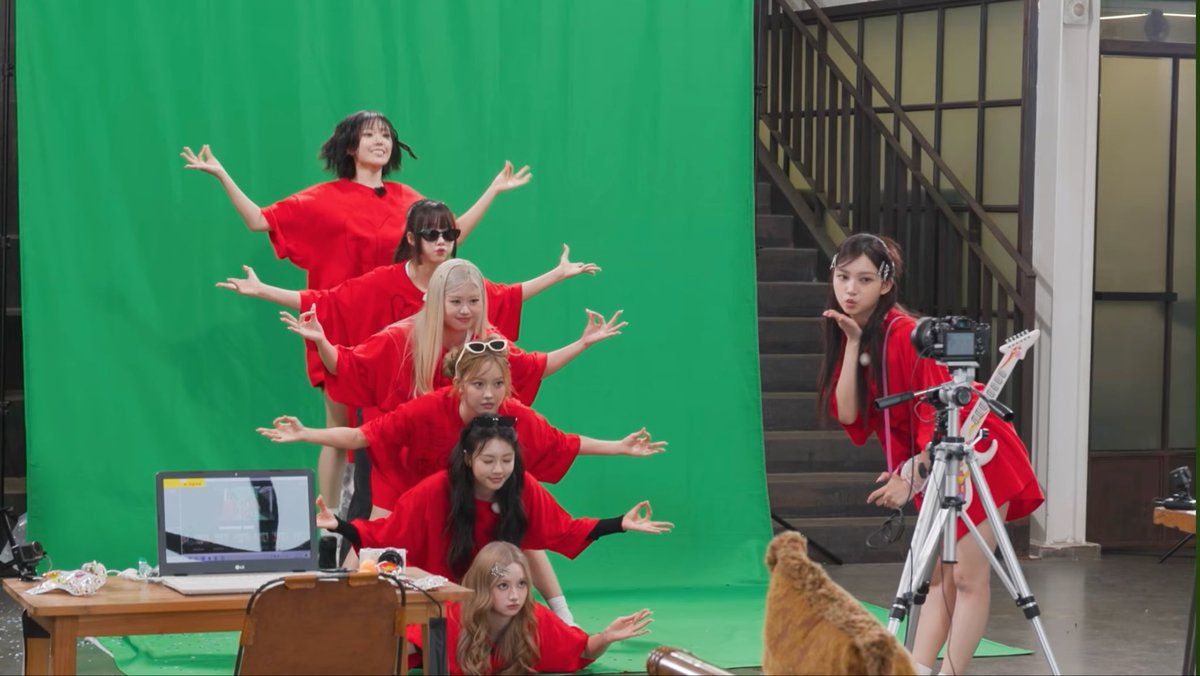 A group of young women in red outfits pose with arms outstretched in front of a green screen, while another stands to the side near a tripod-mounted camera, with a dog visible in the foreground.