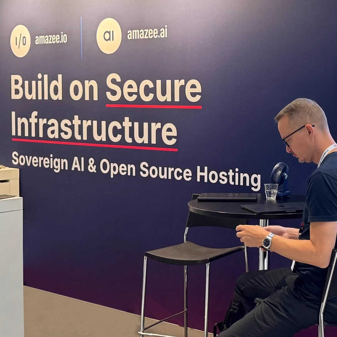 A side-profile view of the amazee.io and amazee.ai booth at DrupalCon Rotterdam 2026. A team member sits at a round table in front of a dark navy backdrop prominently displaying the tagline "Build on Secure Infrastructure: Sovereign AI & Open Source Hosting"