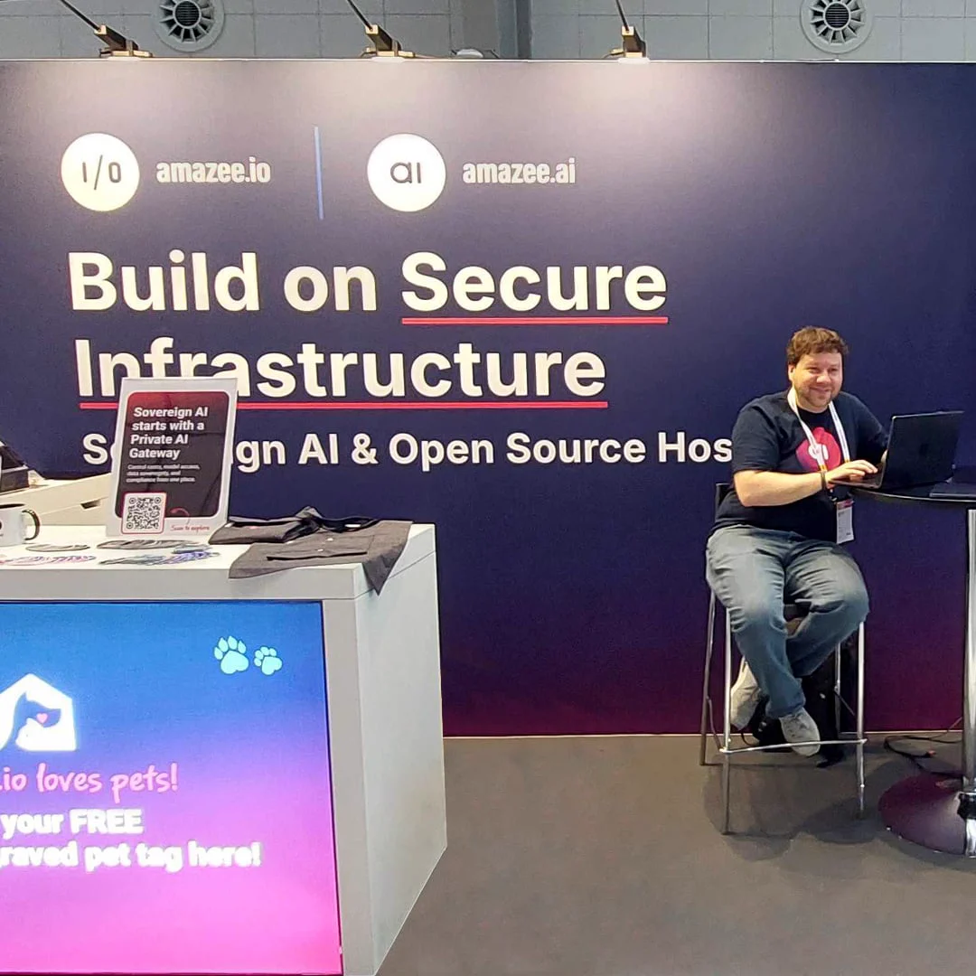 Team member seated at the amazee.io and amazee.ai exhibition booth at DrupalCon Rotterdam 2026, featuring the "Build on Secure Infrastructure" backdrop and free engraved pet tag counter