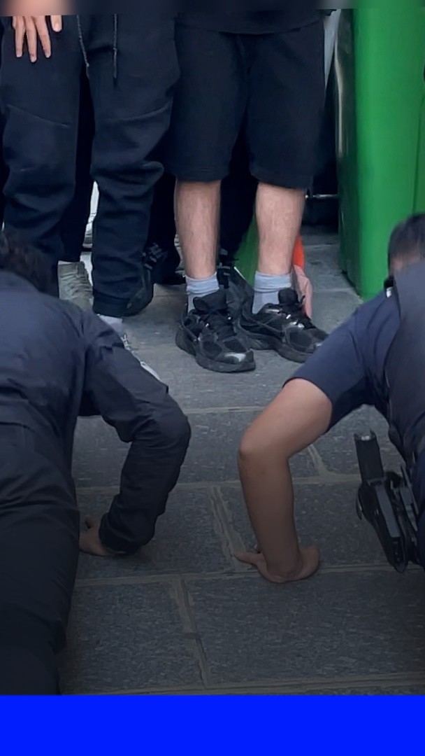A cropped photo showing the lower bodies of several individuals on a paved street; one person is squatting barefoot in handcuffs while a police officer holding a firearm stands nearby.