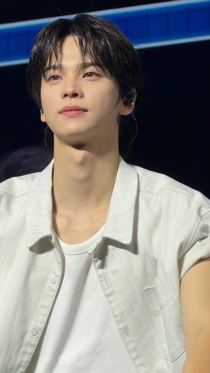 A young man with dark hair wearing a white shirt and in-ear monitors appears to be performing on stage, likely at a K-pop concert.