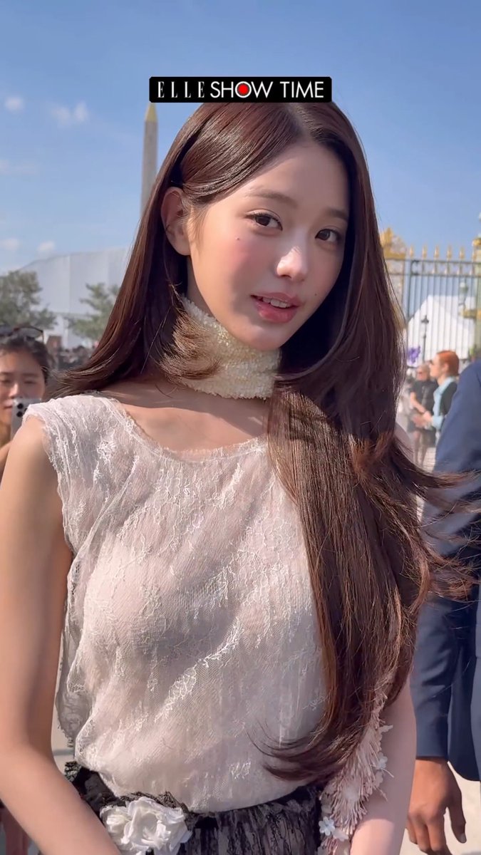 A woman with long brown hair wearing a sheer white lace top and pearl choker poses outdoors, with the text 'ELLE SHOW TIME' displayed above her.