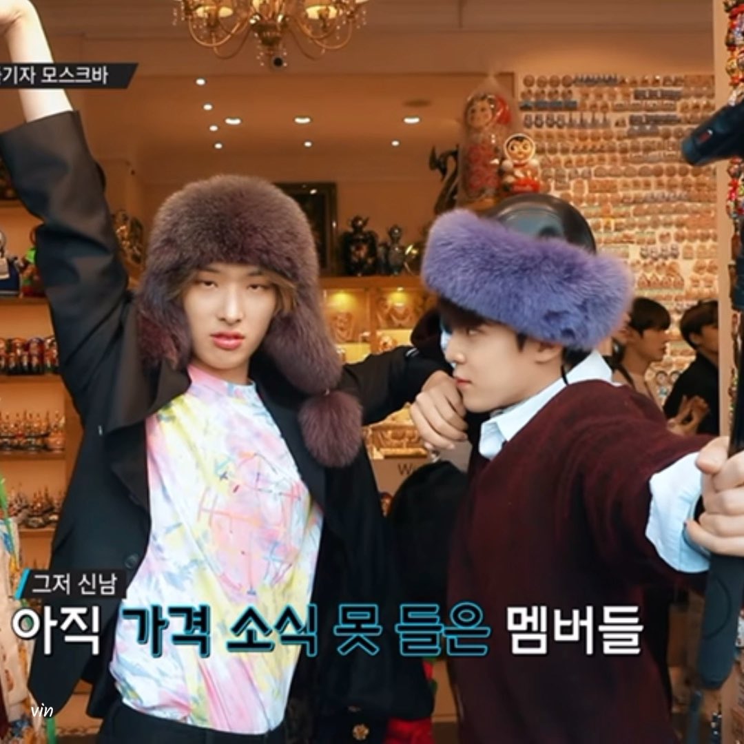 Two young men wearing large furry ushanka hats pose in a souvenir shop filled with Russian nesting dolls, with Korean text overlaid on the image.