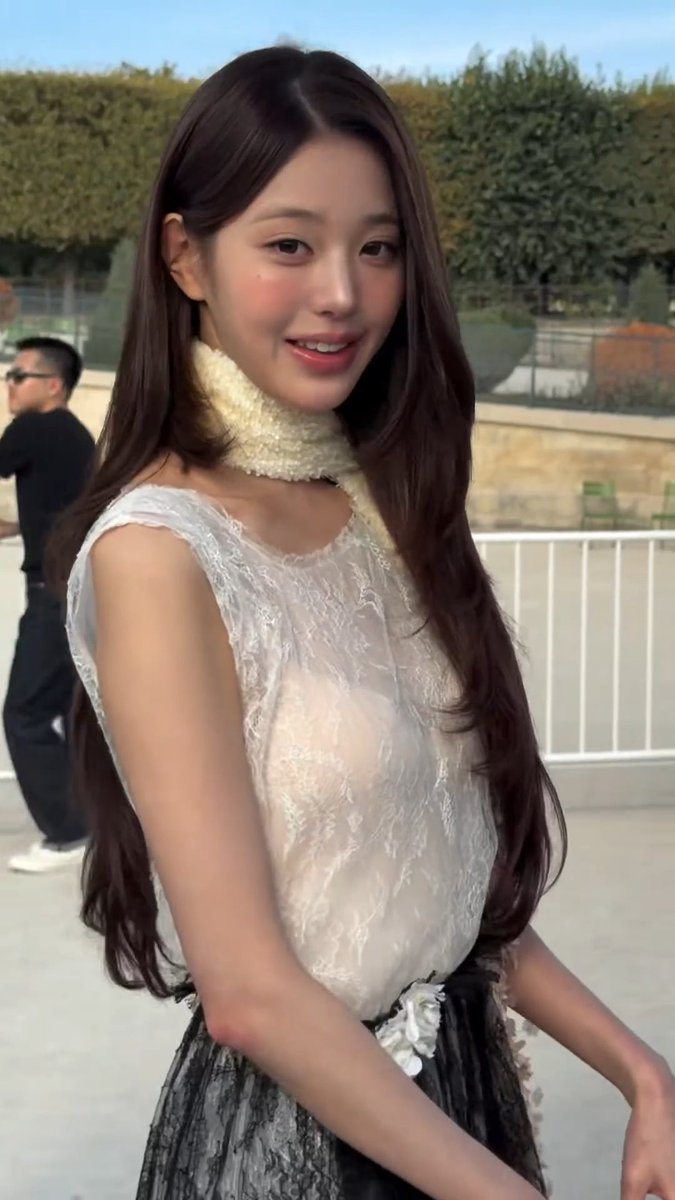 A young woman with long dark hair wears a white lace top and a fluffy scarf, smiling outdoors.