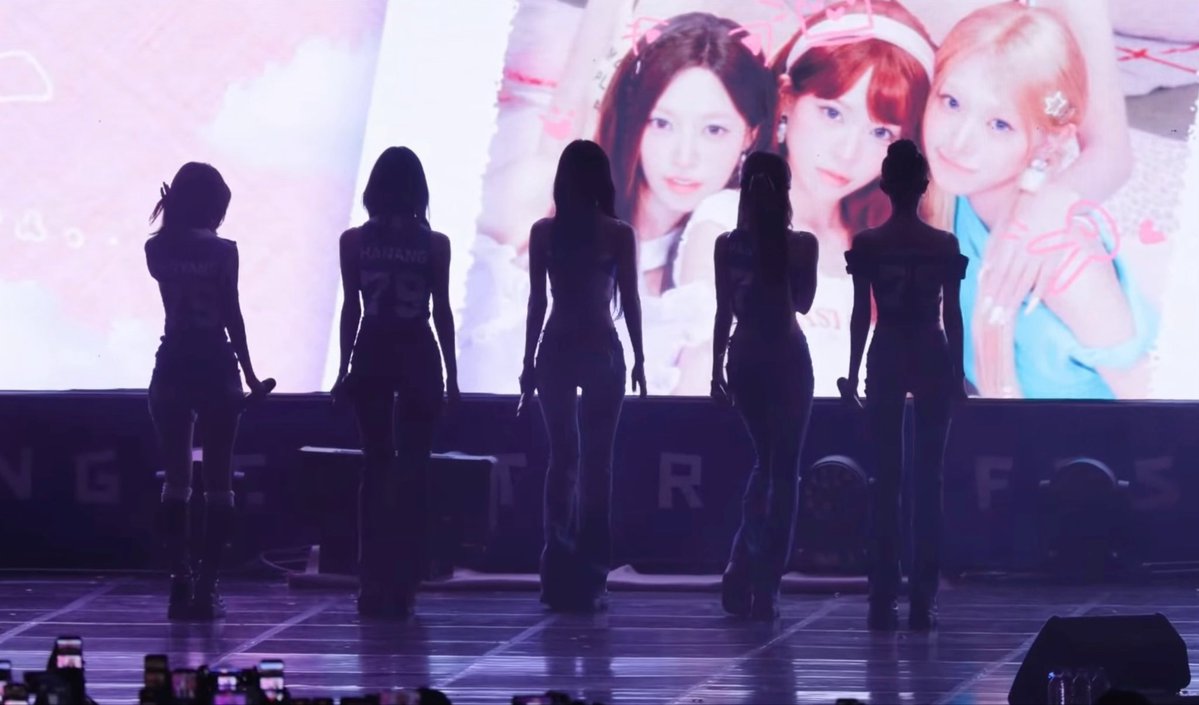 Five silhouetted performers stand on a concert stage with their backs to the camera, wearing form-fitting ripped skinny jeans that emphasize their long, lean silhouettes against the bright LED screen behind them displaying promotional images of the group members. The numbers "79" and partial text "FEST" are visible on the stage backdrop, matching the post's praise for how the outfit's fit dominates even without showing their faces.