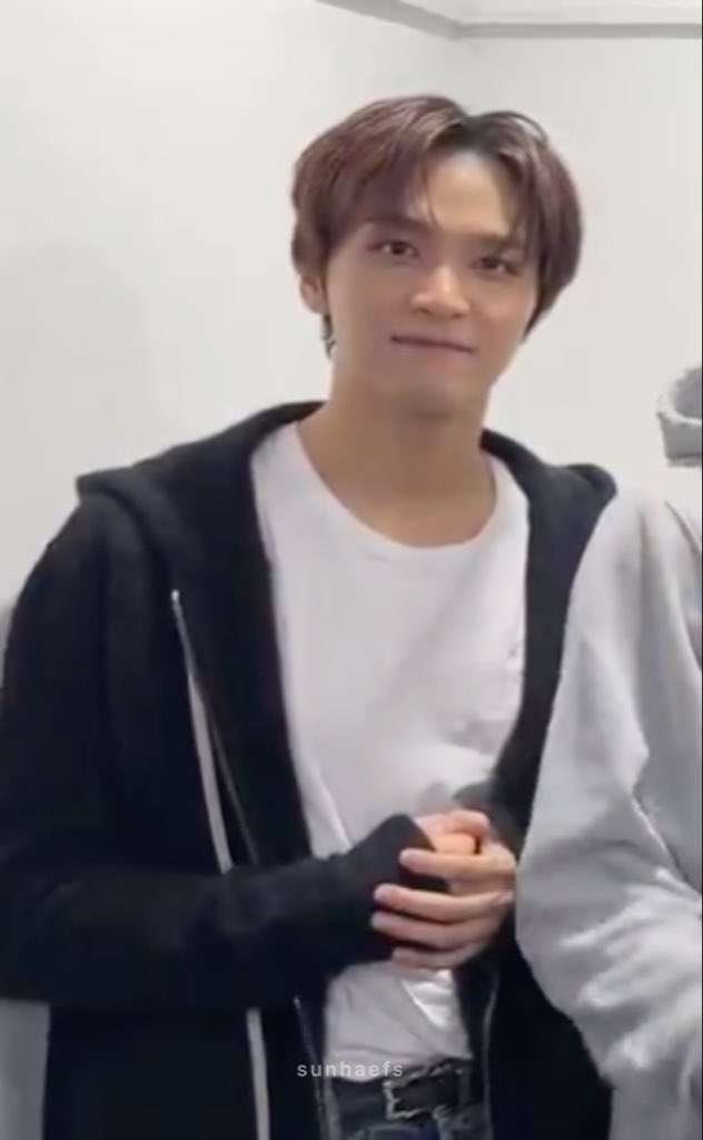 A young man with brown hair, wearing a white t-shirt under a black zip-up hoodie, smiles softly while standing in front of a white background. The watermark "sunhaefs" appears at the bottom, and the caption "see u this weekend haechanie" suggests this is a fan photo of NCT's Haechan looking forward to a weekend event.