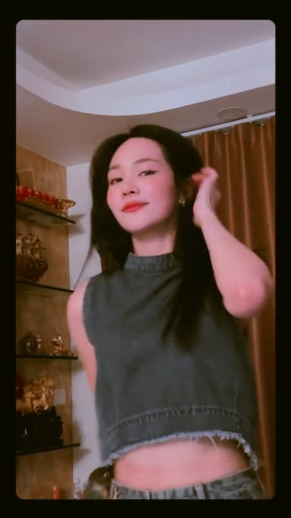A video clip of a young woman with long dark hair wearing a denim crop top, smiling while touching her hair in a room with decorative shelves.