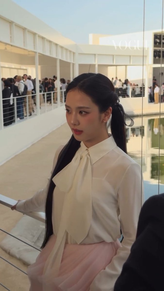 JISOO, the BLACKPINK star and Dior global ambassador, attends the Dior Summer 2027 Women's show in Paris, wearing an elegant ivory blouse with a pussy-bow tie and a soft pink skirt, with "VOGUE" visible on the glass behind her.