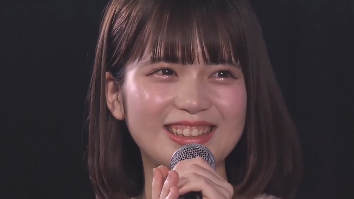 Close-up of a young Japanese woman with brown hair and bangs, smiling with tears on her cheeks while holding a microphone.