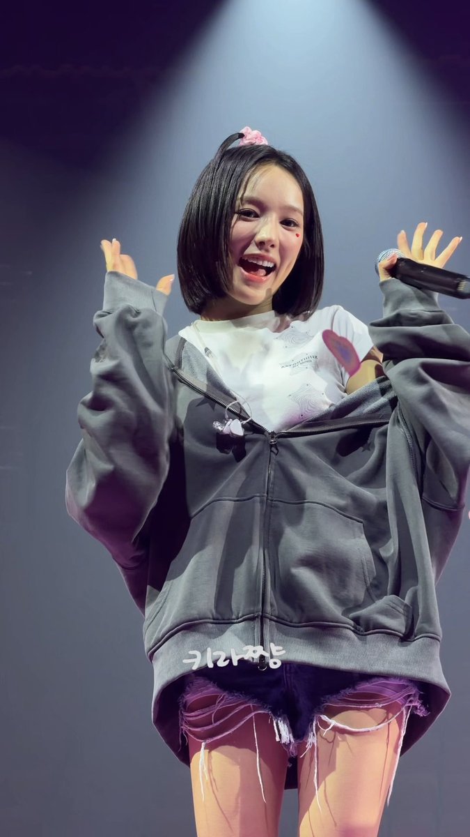 A female performer with short black hair and a pink bow joyfully holds a microphone on stage, wearing an oversized gray zip-up hoodie over a white graphic tee and distressed denim shorts, with Korean text "키라라팡" (Kira Kira) visible on her jacket, matching the post's "sparkle sparkle" theme from #izna's RYUSARANG.