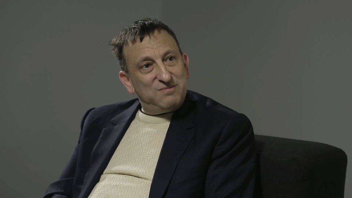 A man with short dark hair, wearing a navy blazer over a cream-colored knit sweater, sits in a black chair against a gray background, appearing engaged in conversation with a slight smile. The image accompanies a post about Brighton chairman Tony Bloom expressing optimism about the club's prospects this season.
