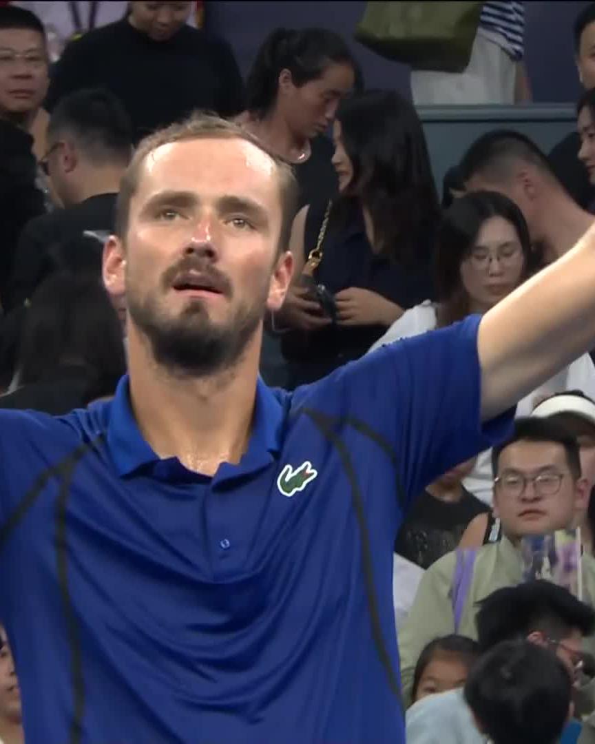 A bearded tennis player wearing a blue Lacoste polo shirt with his arm raised, surrounded by a crowd of spectators.