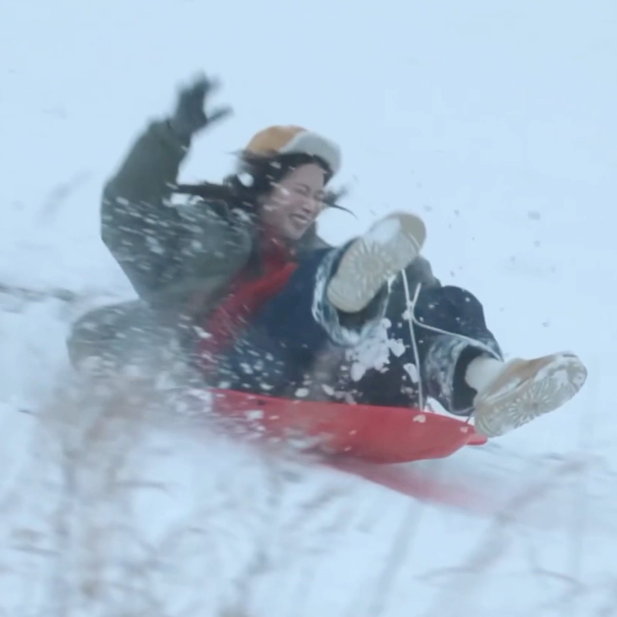 A young woman bundled in winter gear sleds down a snowy hill on a red sled, arms raised in excitement as snow sprays around her, embodying the playful winter fun that connects to the joyful K-pop group LE SSERAFIM and its member Kazuha.
