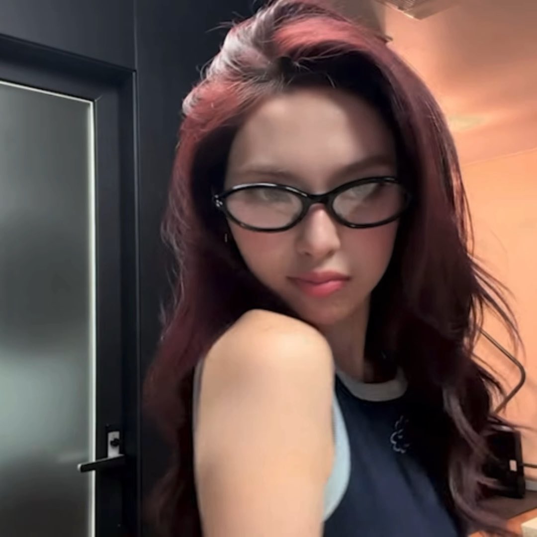 A selfie of a young woman with long reddish-brown hair wearing black-rimmed oval glasses and a dark sleeveless top, posing indoors.