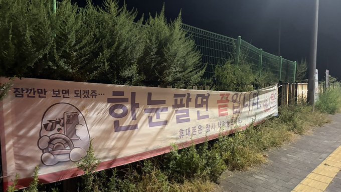 A nighttime photograph of a Korean banner reading '한군 끝엔 쭉익네 (Hangul: end of hangover)' with cartoon character artwork, hung on a fence along a sidewalk.