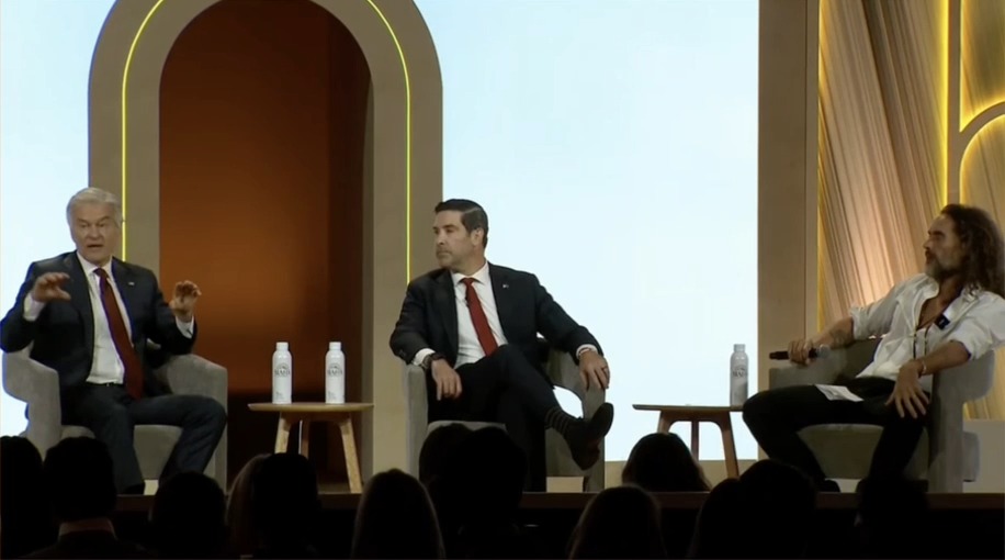 Three men in business attire sit on chairs on a stage during what appears to be a panel discussion, with the man on the left gesturing while speaking. Water bottles with branded labels are placed on small tables between them, and an audience is visible in silhouette in the foreground. The setting suggests a conference or public forum where Dr. Oz is likely participating in the discussion about AI in healthcare referenced in the caption.