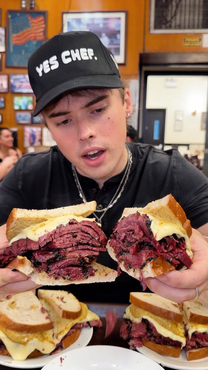 A young man wearing a black 'YES CHEF.' cap holds up two halves of an enormous pastrami sandwich with melted cheese, with additional sandwich portions on plates below.