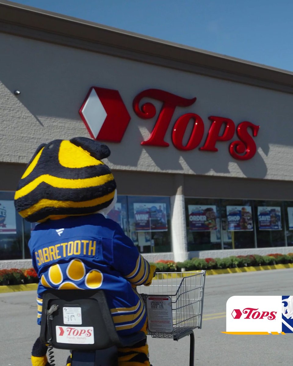 The Buffalo Sabres' mascot Sabretooth, wearing a jersey with his name on the back, pushes a Tops Friendly Markets shopping cart toward a Tops grocery store entrance. The image visually reinforces the team's announcement of Tops Markets continuing as the Official and Exclusive Grocer of the Buffalo Sabres.