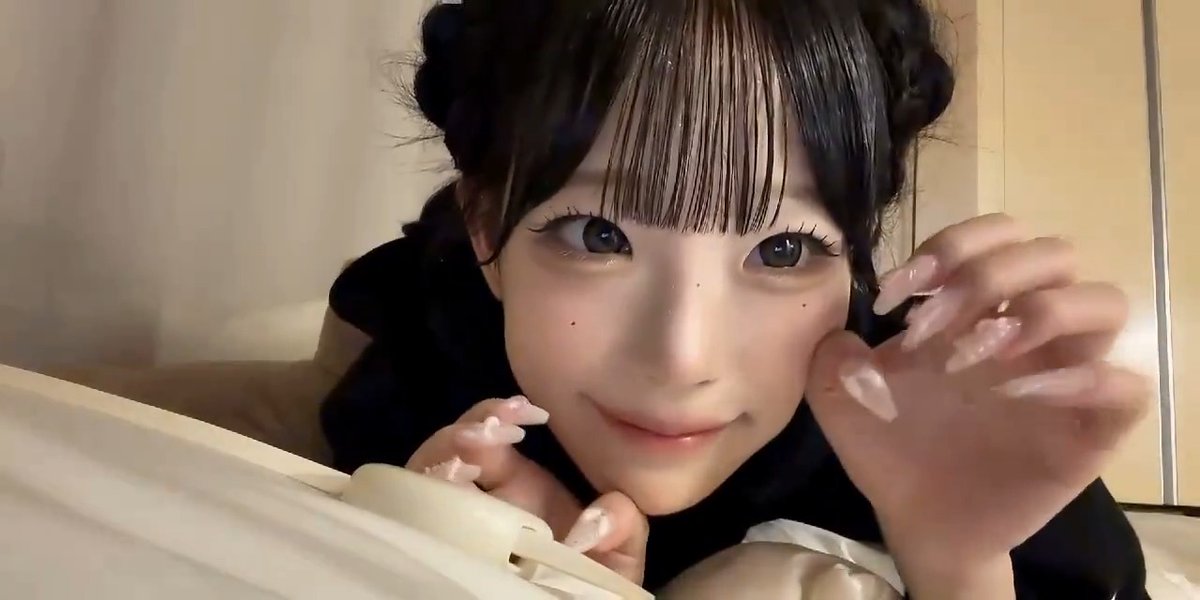 A close-up shot of a person with braided bangs and long, pointed nails smiling softly while leaning on a white ledge.