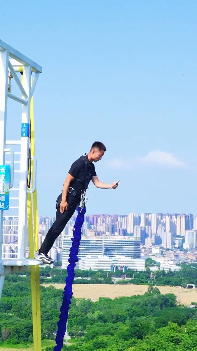 A young man in a black t-shirt prepares to do a bungee jump off a high platform, checking his phone before the leap over a sprawling cityscape.
