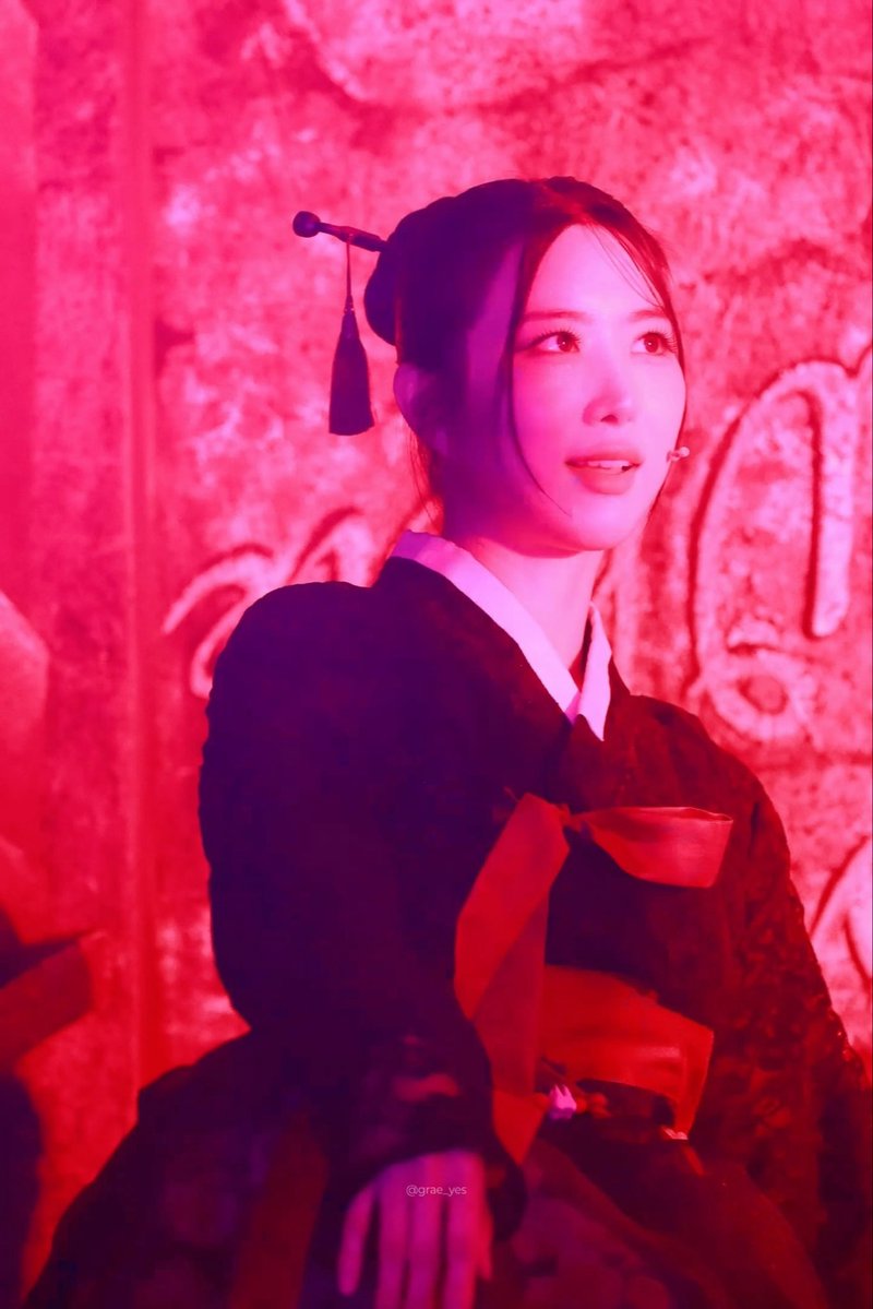 A young woman with dark hair styled in a traditional updo with an ornamental pin performs on stage, bathed in dramatic red lighting, wearing a black and red traditional-style outfit.