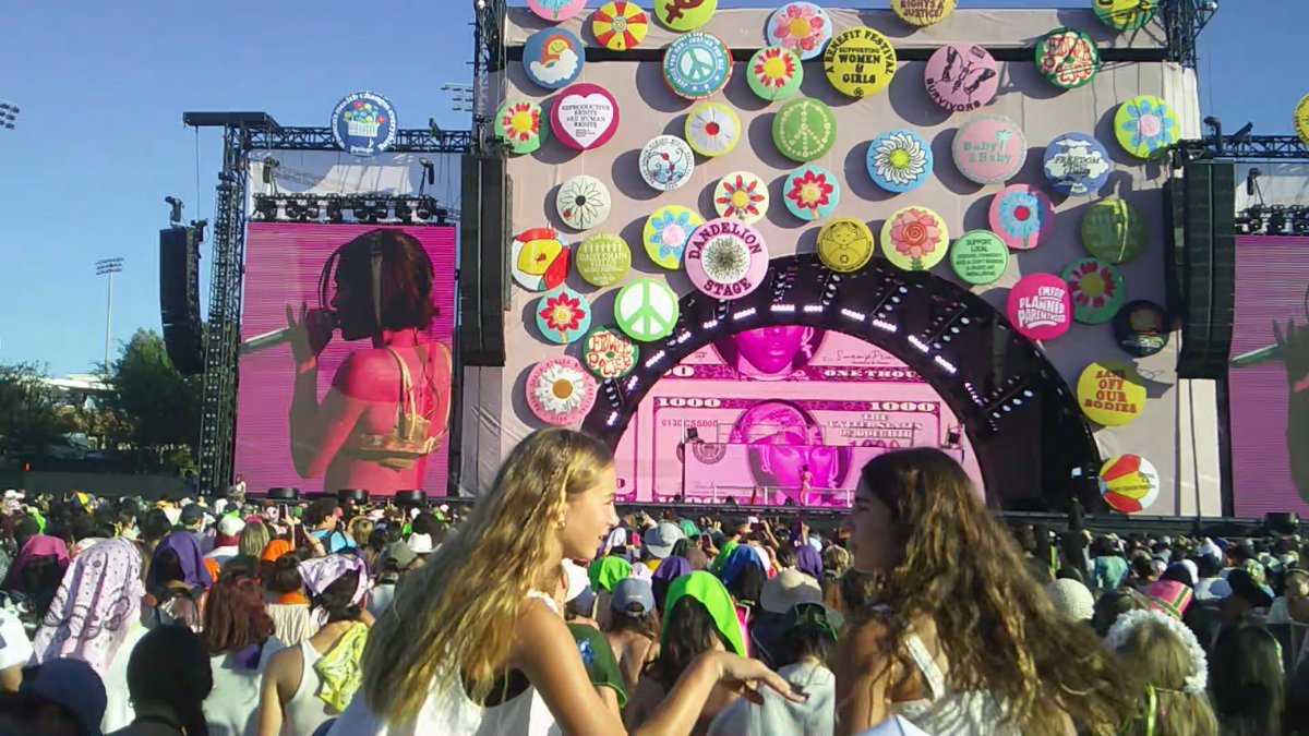 A large crowd attends an outdoor music festival, facing a stage labeled 'DANDELION STAGE' decorated with numerous colorful, flower-themed circular signs.