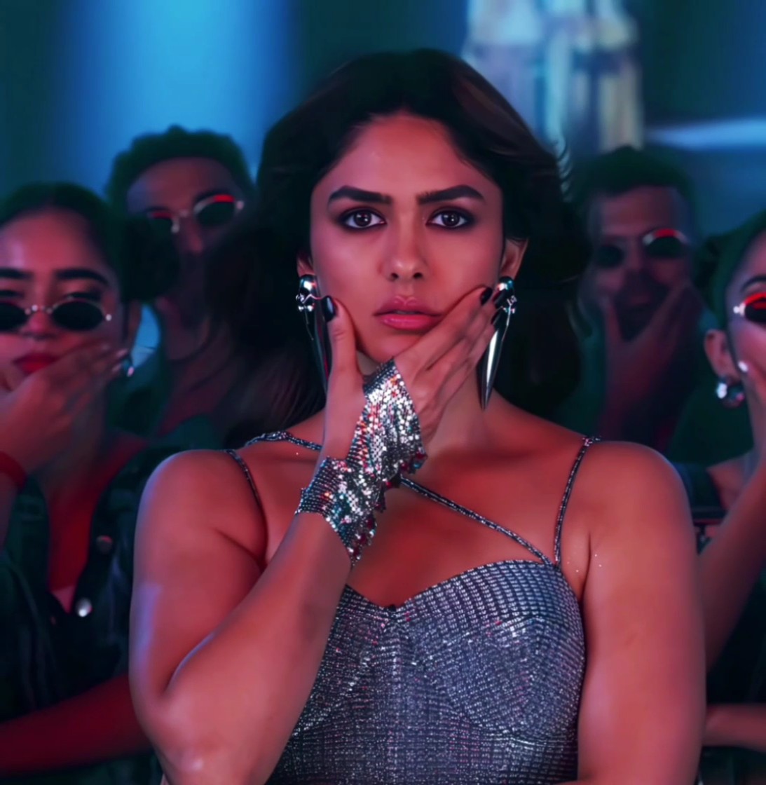 A glamorous woman with dramatic makeup and a sparkly silver dress strikes a pose with her hand on her face, surrounded by backup dancers wearing sunglasses in a club-like setting with blue and pink lighting.